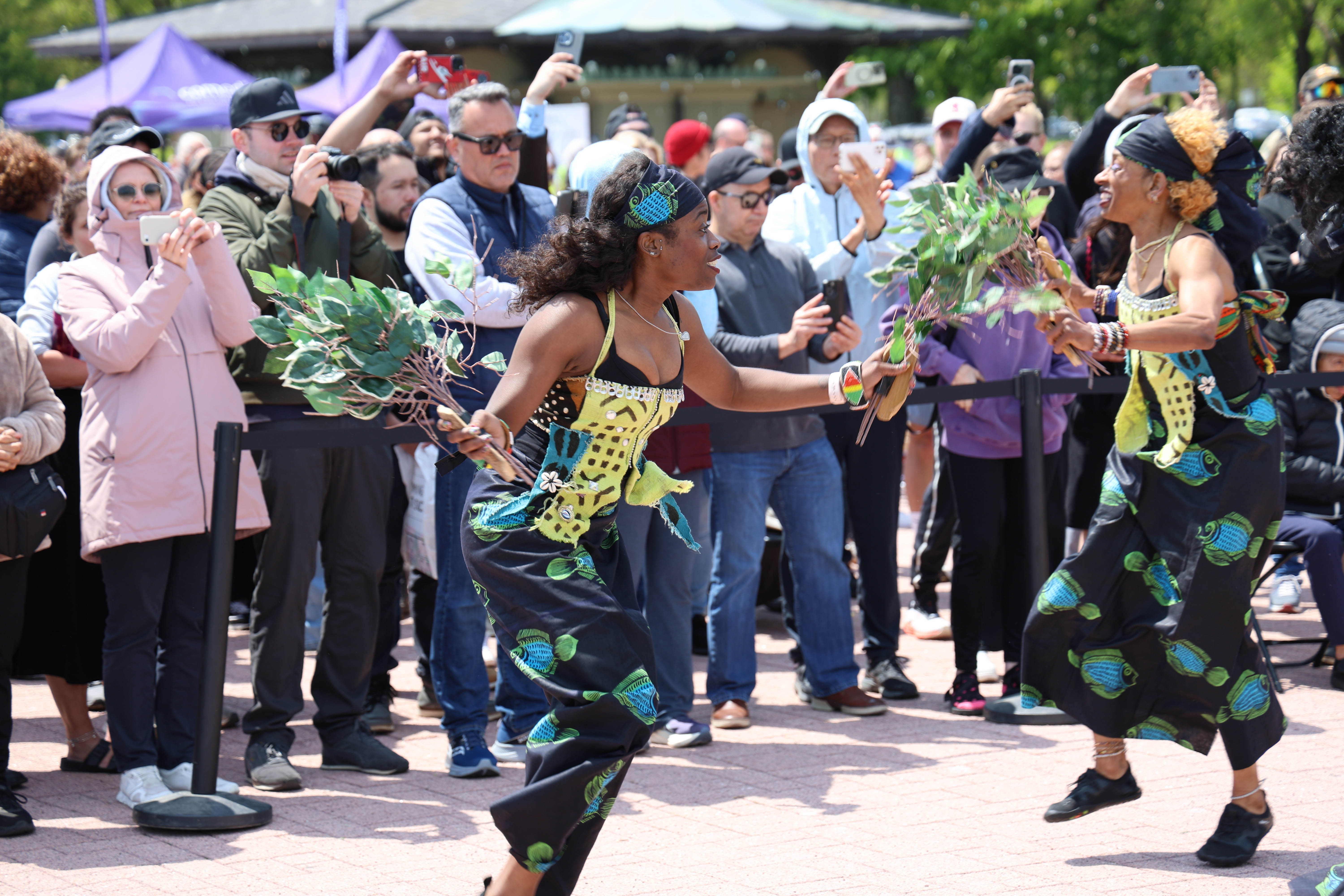 Two women in colorful patterned clothing dance, holding branches, as onlookers take photos.